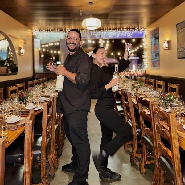 Two Agave staff members, back-to-back, holding bottles of spirits in a festive dining room.