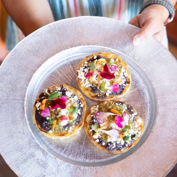 Three Mexican sopes with black beans, green sauce, cheese, and edible flowers on a clear plate.