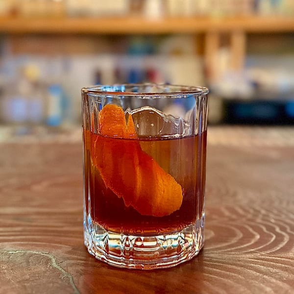 A dark cocktail with an orange peel garnish and ice in a rocks glass on a wooden table.