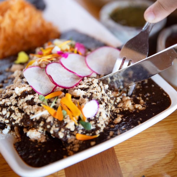 A person cutting into a Mexican dish with dark sauce, crumbled cheese, radishes, and edible flowers.