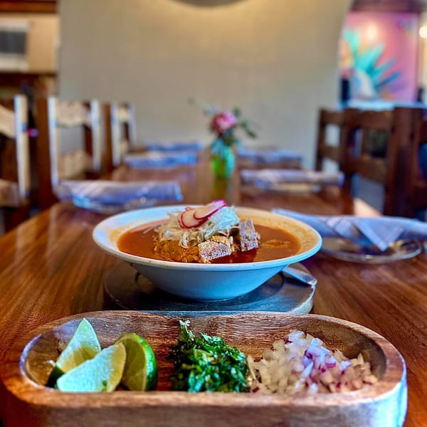 A bowl of Agave's Mexican pozole with radish, cabbage, and meat, served with lime, cilantro, and onions.