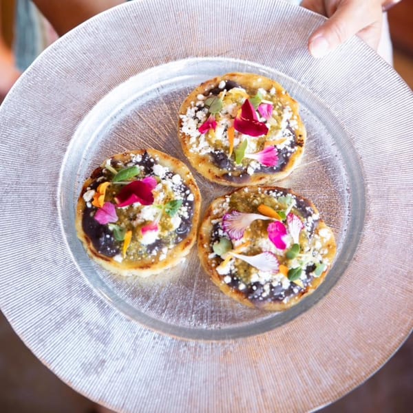 Three Mexican sopes with black beans, cheese, green sauce, and edible flowers on a silver plate.