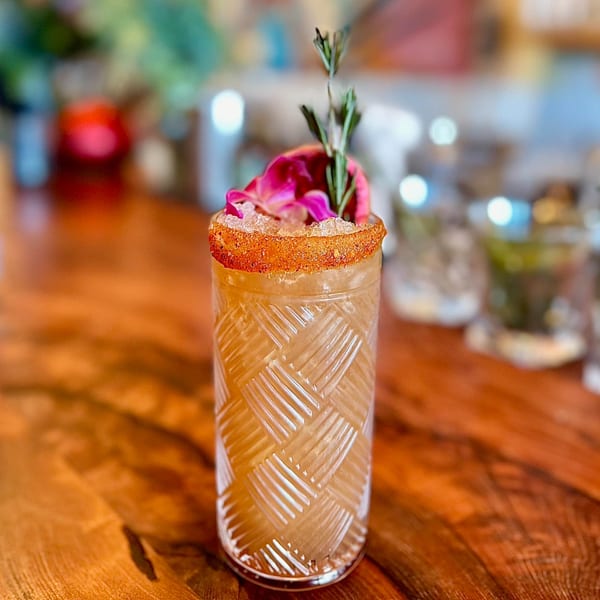 A tall, patterned glass of Agave's Mexican cocktail, garnished with rosemary and pink flower petals.