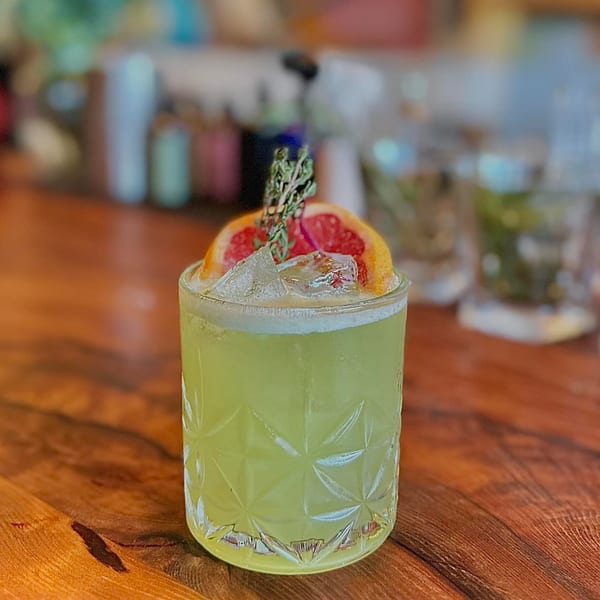 A green cocktail with ice, grapefruit slices, and a sprig of thyme on a wooden table.