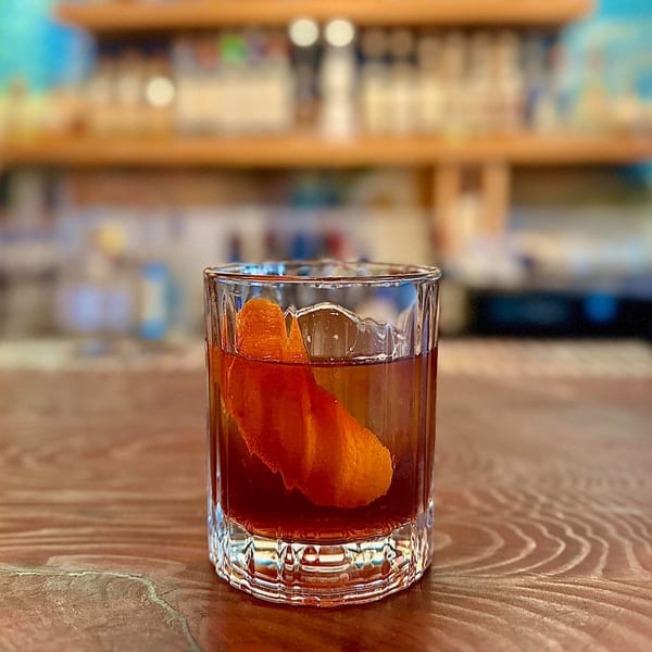 A dark cocktail with an orange peel garnish sits on a wooden bar at Agave.