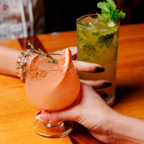 Two hands holding a pink cocktail with a grapefruit slice and a green mojito on a wooden table.