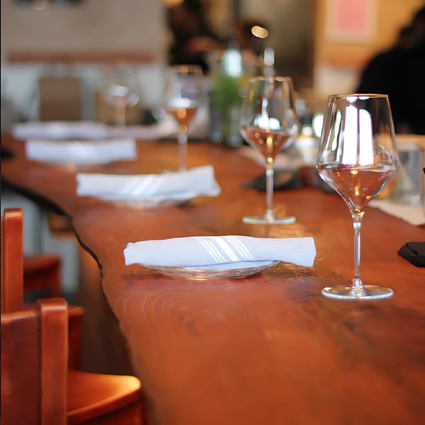 Set tables with wine glasses and folded napkins on a long, polished wooden bar at Agave.