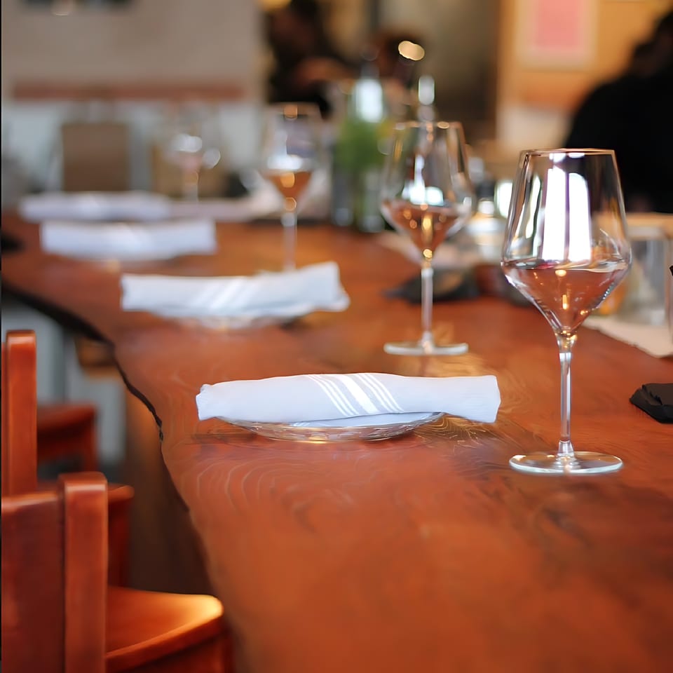 Set tables with wine glasses and folded napkins on a long, polished wooden bar at Agave.