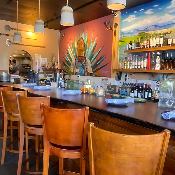 Agave Mexican Restaurant's bar with wooden stools, a colorful mural, and bottles of spirits.