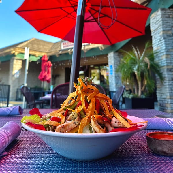 A vibrant Mexican salad with chicken, tortilla strips, and peppers, under a red umbrella on a patio.