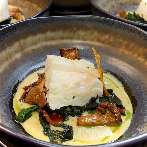 White fish with mushrooms and greens in a creamy sauce, served in a dark bowl.