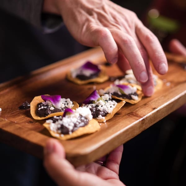 Hands presenting a wooden tray of Agave's Mexican appetizers with black beans, cheese, and purple flowers.
