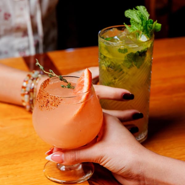 Two hands holding cocktails, one a pink grapefruit drink, the other a green mojito.