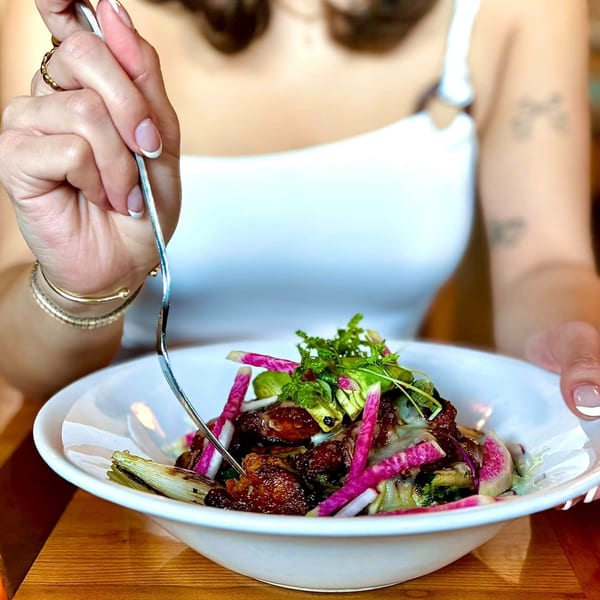 A person in a white top uses a fork to eat a Mexican dish with meat, avocado, and radishes.