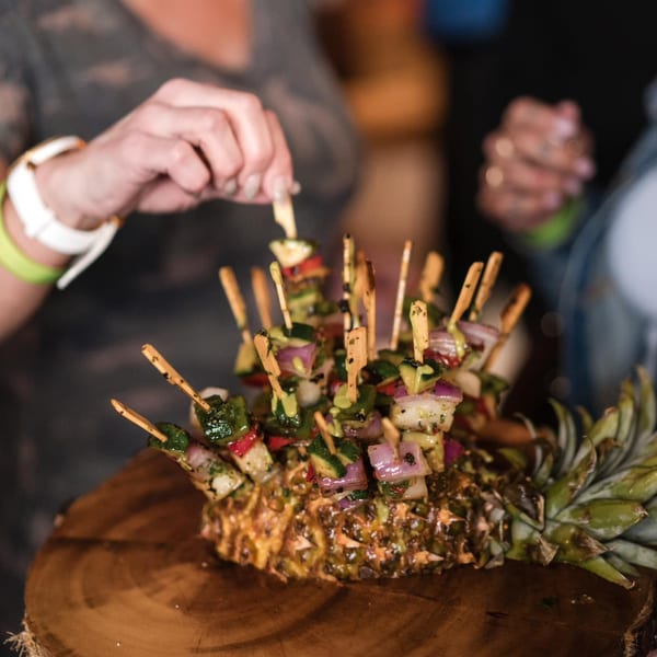 A person taking a skewer of grilled vegetables and shrimp from a pineapple display.